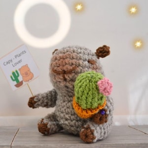 Puede incluir: Un peluche de capibara hecho a mano con ganchillo en tonos grises, marrones, verdes y naranjas. El peluche sostiene un cactus y un letrero que dice "Capy - Plants Lover". El peluche está sentado sobre una superficie de madera.