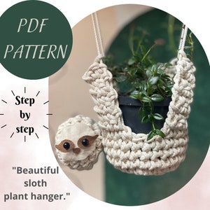 May include: A crocheted sloth plant hanger made with beige rope and a small brown sloth face. The hanger is holding a potted plant with green leaves.
