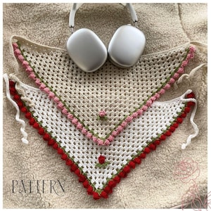 Puede incluir: Dos bufandas triangulares de crochet con fondo blanco y detalles en rojo, rosa y verde. Las bufandas están superpuestas y tienen un borde de flecos. Las bufandas están sobre una superficie blanca peluda.