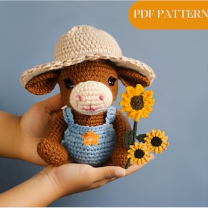 Könnte beinhalten: Eine gehäkelte braune Kuh, die einen Strohhut und eine blaue Latzhose trägt und eine Sonnenblume mit grünen Blättern hält.