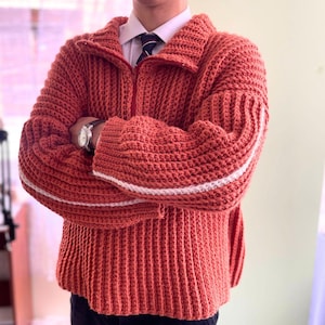 Puede incluir: Un suéter de punto rojo con rayas blancas en las mangas. El suéter tiene una cremallera y un cuello alto.