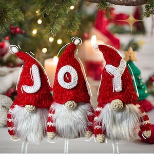 Puede incluir: Tres gnomos de crochet rojo con barbas de piel blanca y letras de fieltro blanco que deletrean "JOY" en sus sombreros. Los gnomos están de pie frente a un árbol de Navidad y luces.