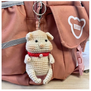 May include: A crocheted dog keychain with a red collar and a pearl accent. The dog is beige with black eyes and a brown nose. The keychain is attached to a pink backpack with the word "LEISURE" printed on it.