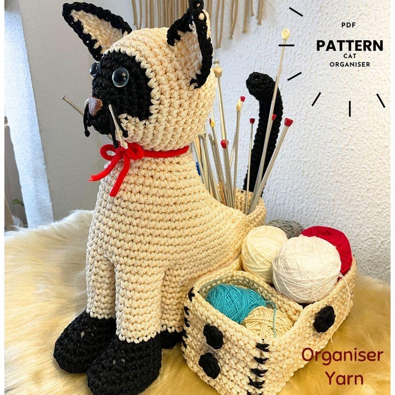 May include: A crocheted Siamese cat-shaped organizer with black and cream yarn. The cat has a red collar and holds knitting needles. A basket with yarn balls sits beside it. Text on the image reads "Pattern Cat Organiser Yarn".