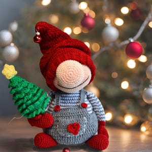 Puede incluir: Un gnomo de crochet con un gorro rojo y sosteniendo un pequeño árbol de Navidad verde. El gnomo está sentado en una superficie de madera con un fondo borroso de luces navideñas.
