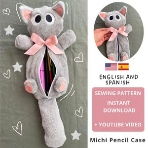 Puede incluir: Un estuche para lápices con forma de gato gris de felpa, con un lazo rosa y almohadillas rosas. El estuche está abierto, revelando bolígrafos y lápices de colores en el interior. La imagen contiene texto.