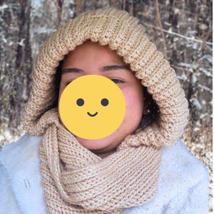 Puede incluir: Conjunto de gorro y bufanda de punto beige. El gorro está diseñado con un patrón de canalé y está conectado a una bufanda a juego. El conjunto está hecho de un material suave y cálido, perfecto para el frío. El fondo es una escena exterior borrosa.