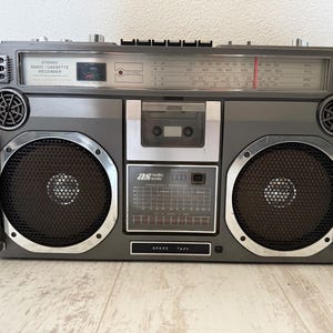 Może przedstawiać: Vintage szary magnetofon stereo z radiem i kasetą ze srebrnymi akcentami. Boombox posiada duże głośniki, gniazdo na kasetę i pokrętło radia. Widoczny jest tekst "STEREO RADIO/CASSETTE RECORDER".