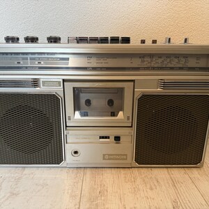 May include: Silver vintage Hitachi boombox with a cassette player, radio dial, and two black speaker grilles. The boombox has several knobs and buttons, and the Hitachi logo is visible.