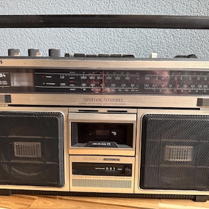 Op de afbeelding: Een zilverkleurige en zwarte Philips 584 Spatial Stereo 4-bands AM/FM radiocassetterecorder. Het apparaat is voorzien van een handvat aan de bovenzijde, een centraal cassettedeck, twee grote zwarte luidsprekerroosters en een horizontale zenderzoeker.