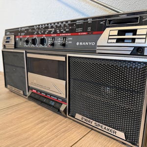 Op de afbeelding: Een vintage Sanyo boombox met een zilver en zwart kleurenschema. De radio heeft een cassettespeler, afstemknop en luidsprekerroosters. De tekst "SANYO" is zichtbaar op de voorkant. De boombox heeft een telescopische antenne en diverse bedieningsknoppen.