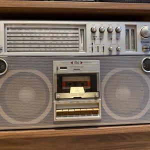 Op de afbeelding: Een zilverkleurige draagbare radiocassettespeler met een groot radiofrequentiedisplay, meerdere bedieningsknoppen, twee grote ronde luidsprekers en een centraal cassettedeck met een cassette erin. De merknaam SILVER staat op de voorzijde gedrukt.