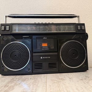 May include: Black Sanyo boombox with silver detailing, including a cassette player and radio dial. The boombox has two large speakers and a handle. The Sanyo logo is visible on the front.