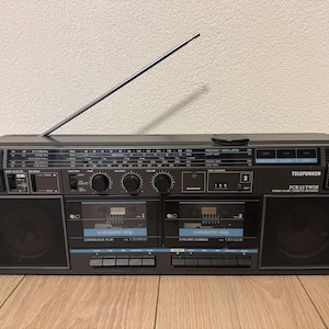 Könnte beinhalten: Ein schwarzer Telefunken PCR 33 Twin Boombox aus den 80er Jahren mit silberner Antenne. Das Stereokassetten-Aufnahmegerät hat zwei Kassettenfächer, Lautsprecher und verschiedene Knöpfe. Die Boombox steht auf einer Holzoberfläche.