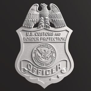May include: A silver-toned U.S. Customs and Border Protection officer badge featuring an eagle at the top, the text U.S. CUSTOMS AND BORDER PROTECTION, a central Department of Homeland Security seal, and the word OFFICER on a banner at the bottom.