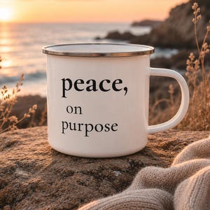 Könnte beinhalten: Ein weißer Emaillebecher mit silbernem Rand und dem schwarzen Text "peace, on purpose" auf der Vorderseite. Der Becher hat einen geschwungenen Henkel auf der rechten Seite und steht auf einer strukturierten Felsoberfläche.