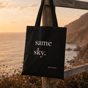 Könnte beinhalten: Eine schwarze Tragetasche mit dem weißen Schriftzug same sky. in Serifenschrift in der Mitte und dem kleineren weißen Text soft cosmos unten rechts. Die Tasche hat zwei lange Schulterriemen und ein kleines grünes Stoffetikett an der Seite.