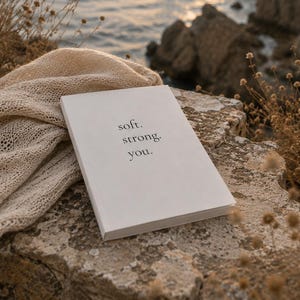 Könnte beinhalten: Ein weißes Buch mit dem schwarzen Aufdruck soft. strong. you. auf dem Einband, begleitet von einem beigen Strickstoff, der daneben liegt. Das Buch befindet sich auf einer strukturierten Steinoberfläche mit einer Küstenlandschaft im Hintergrund.