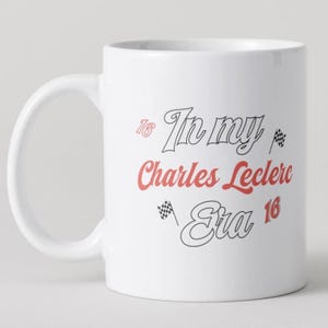 May include: A white ceramic mug featuring black and red text that reads In my Charles Leclerc Era. The design includes the number 16 in red and black, along with two small black and white checkered racing flags positioned near the text.