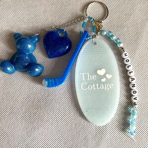 May include: A keychain featuring a blue glittery bear charm, a dark blue glittery heart, a blue hockey stick, and an oval-shaped light blue tag with the text The Cottage and three small hearts. A string of blue and white beads spells HOLLANOV.