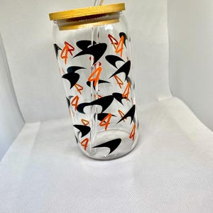 K&ouml;nnte beinhalten: Ein durchsichtiger Glastrinkbecher mit Bambusdeckel und Glasstrohhalm, der mit einem sich wiederholenden Muster aus schwarzen abstrakten Formen und der orangefarbenen Zahl 4 auf der Au&szlig;enseite des zylindrischen Gef&auml;&szlig;es bedruckt ist.