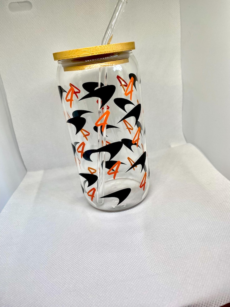 K&ouml;nnte beinhalten: Ein durchsichtiger Glastrinkbecher mit Bambusdeckel und Glasstrohhalm, der mit einem sich wiederholenden Muster aus schwarzen abstrakten Formen und der orangefarbenen Zahl 4 auf der Au&szlig;enseite des zylindrischen Gef&auml;&szlig;es bedruckt ist.