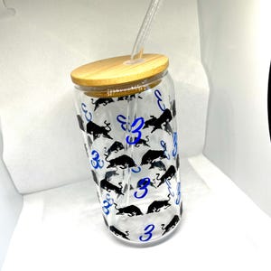 K&ouml;nnte beinhalten: Ein durchsichtiges Glas mit Bambusdeckel und Glasstrohhalm. Die Au&szlig;enseite ist mit einem sich wiederholenden Muster aus schwarzen Stiersilhouetten und der blauen Zahl 3 bedruckt, die &uuml;ber die Oberfl&auml;che des zylindrischen Gef&auml;&szlig;es verteilt sind.