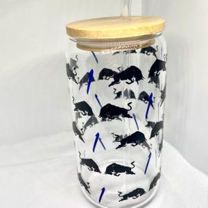 K&ouml;nnte beinhalten: Ein durchsichtiger Glasbecher mit Bambusdeckel und Glasstrohhalm. Die Au&szlig;enseite des Glases ist mit einem sich wiederholenden Muster aus schwarzen Stiersilhouetten und kleinen, d&uuml;nnen blauen vertikalen Linien &uuml;ber die zylindrische Oberfl&auml;che verziert.