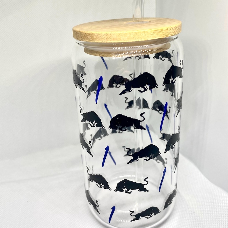 K&ouml;nnte beinhalten: Ein durchsichtiger Glasbecher mit Bambusdeckel und Glasstrohhalm. Die Au&szlig;enseite des Glases ist mit einem sich wiederholenden Muster aus schwarzen Stiersilhouetten und kleinen, d&uuml;nnen blauen vertikalen Linien &uuml;ber die zylindrische Oberfl&auml;che verziert.