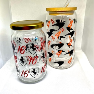 K&ouml;nnte beinhalten: Zwei klare Glastrinkbecher mit Deckeln und einem Strohhalm. Das k&uuml;rzere Glas zeigt rote Ziffern 16 und schwarze Pferdeschild-Grafiken. Das h&ouml;here Glas zeigt orangefarbene Ziffern 4 und schwarze Schwung-Grafiken. Beide Gl&auml;ser haben goldene Deckel.