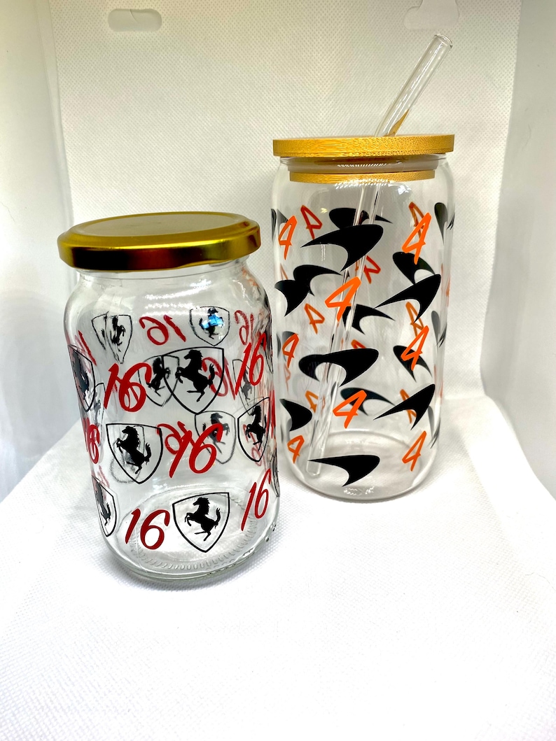 K&ouml;nnte beinhalten: Zwei klare Glastrinkbecher mit Deckeln und einem Strohhalm. Das k&uuml;rzere Glas zeigt rote Ziffern 16 und schwarze Pferdeschild-Grafiken. Das h&ouml;here Glas zeigt orangefarbene Ziffern 4 und schwarze Schwung-Grafiken. Beide Gl&auml;ser haben goldene Deckel.