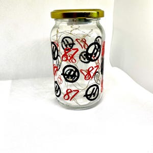 K&ouml;nnte beinhalten: Ein klares Glasgef&auml;&szlig; mit einem goldfarbenen Metalldeckel, verziert mit einem sich wiederholenden Muster aus der roten Zahl 87 und einem schwarzen kreisf&ouml;rmigen Logo. Im Inneren des Glases sind d&uuml;nne, kupferfarbene Lichterketten aufgewickelt.