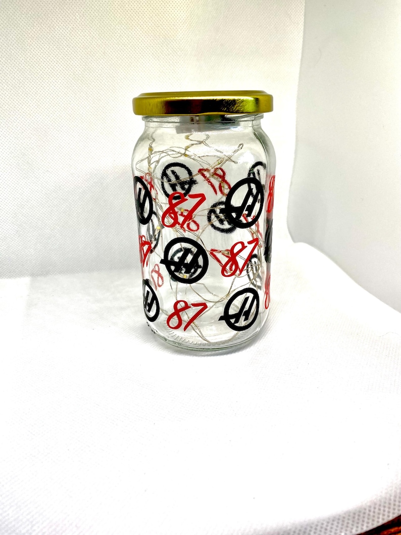K&ouml;nnte beinhalten: Ein klares Glasgef&auml;&szlig; mit einem goldfarbenen Metalldeckel, verziert mit einem sich wiederholenden Muster aus der roten Zahl 87 und einem schwarzen kreisf&ouml;rmigen Logo. Im Inneren des Glases sind d&uuml;nne, kupferfarbene Lichterketten aufgewickelt.