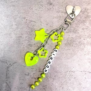 Könnte beinhalten: Ein Schlüsselanhänger mit einem silberfarbenen herzförmigen Verschluss und Ring. Er enthält neon-gelbe Anhänger in Form eines massiven Sterns, eines Herzens und zwei Sternumrissen. Eine Kette aus lindgrünen Perlen bildet den Namen NORRIS in weißen Buchstaben.