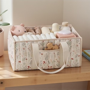 Peut inclure: Un organisateur pour bébé à motifs floraux rempli de couches, de jouets et d'articles de toilette. Le bac en tissu a une poche avant et des poignées. Une plante en pot rose et un livre pour enfants sont à proximité.