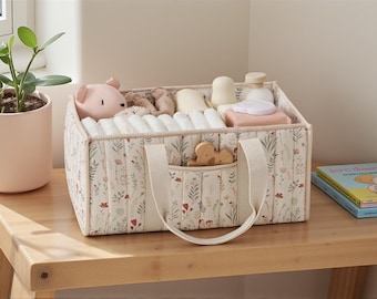 Op de afbeelding: Een bloemmotief baby-organizer gevuld met luiers, speelgoed en toiletartikelen. De stoffen bak heeft een voorvak en handvatten. Een roze potplant en een kinderboek staan ​​in de buurt op een houten tafel.