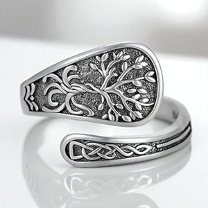 Puede incluir: Anillo plateado con un diseño de árbol de la vida en la parte superior. El anillo tiene una banda ajustable con un diseño de nudo celta. El árbol de la vida está detallado con hojas y ramas. El anillo es probablemente de metal.
