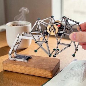 Op de afbeelding: Een zilverkleurig, geometrisch spinnenvormig mechanisch model met een houten basis. Het model heeft gelede poten met groene accenten en een centraal mechanisme met gouden en zwarte componenten. Een hand houdt het model vast, met een koffiekopje en een boek op de achtergrond.