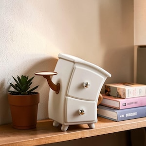 Puede incluir: Una mesita de noche blanca caprichosa con dos cajones y un diseño curvo. Cuenta con pomos dorados y una pequeña estantería adjunta. Una suculenta en maceta está a la izquierda, y los libros están apilados sobre una superficie de madera.