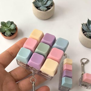 May include: Pastel-coloured keyboard keychains. The keycaps are in shades of yellow, pink, green, purple, and blue. The keychains are attached to a silver chain and a clear plastic base. The background includes small potted succulents.