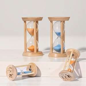 Puede incluir: Cuatro relojes de arena de madera con bombillas de vidrio transparente. Dos relojes de arena contienen arena naranja, uno tiene arena azul y otro tiene arena azul y naranja. Los marcos de madera son de color marrón claro. Un reloj de arena tiene un diseño de dibujos animados y el texto "1 min".
