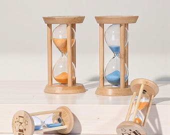 Op de afbeelding: Vier houten zandlopers met heldere glazen bollen. Twee zandlopers bevatten oranje zand, één heeft blauw zand en één heeft blauw en oranje zand. De houten frames zijn lichtbruin. Eén zandloper heeft een cartoonontwerp en de tekst "1 min".