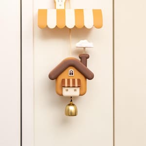 Puede incluir: Un carillón de viento decorativo con forma de casa. La casa es de color naranja con tejado y chimenea marrones, una pequeña ventana y una campana de latón. Cuelga de un toldo a rayas y una cuerda.