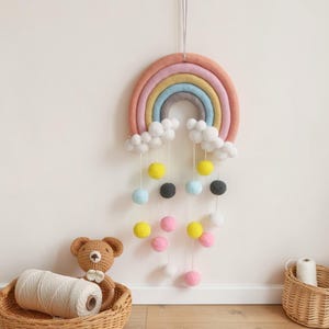 Puede incluir: Un adorno de pared de fieltro con forma de arcoíris en colores pastel, con bolas de fieltro colgantes de varios colores. El arcoíris tiene capas de rosa, amarillo, azul y gris. Nubes blancas de fieltro están unidas.