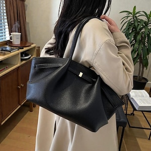 Puede incluir: Un bolso tote de cuero negro con un acabado texturizado. El bolso tiene dos asas y un cierre dorado. El bolso se lleva sobre el hombro. El fondo incluye un mueble de madera y una planta.