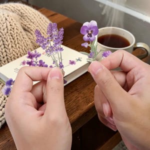 Puede incluir: Primer plano de una persona sosteniendo dos ramitas de flores moradas, una de lavanda y otra de pensamiento. Una taza de café está en el fondo, junto con un cuaderno y una manta de punto beige.