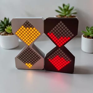May include: Two digital hourglass clocks. One is white with yellow lights, the other is dark brown with red lights. Each clock has a grid of lights in a diamond shape. The clocks are on a white surface with plants in the background.