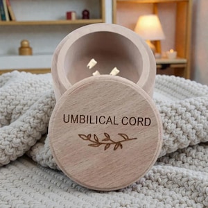Puede incluir: Una caja de recuerdo de madera de color claro con las palabras "UMBILICAL CORD" grabadas en la tapa. La caja está abierta, revelando pequeños objetos blancos en el interior. Una ramita decorativa está grabada debajo.