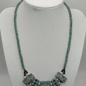 Op de afbeelding: Een kralenketting met een turquoise en bruin geometrisch ontwerp. De ketting heeft een geweven turquoise ketting en rechthoekige hangers met witte en bruine bloempatronen. Zwarte en witte kralen accentueren het ontwerp.