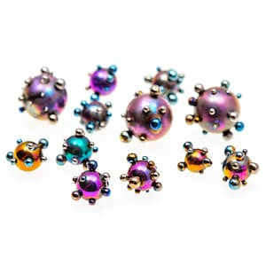 Puede incluir: Una colección de cuentas redondas iridiscentes multicolores con una superficie texturizada. Las cuentas están dispuestas sobre un fondo blanco.